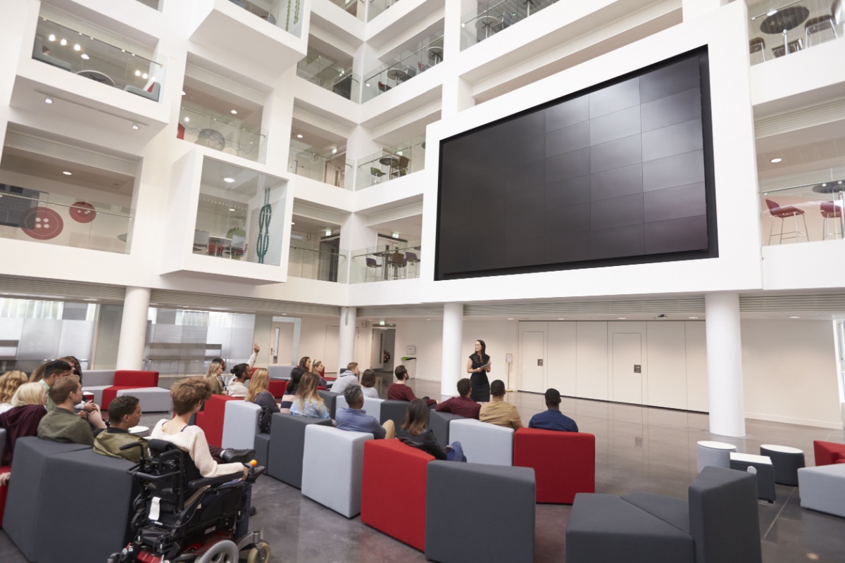 Student lecture in atrium of modern university with large screen.